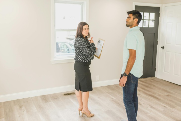 two people looking around a home with white walls and wooden floor.