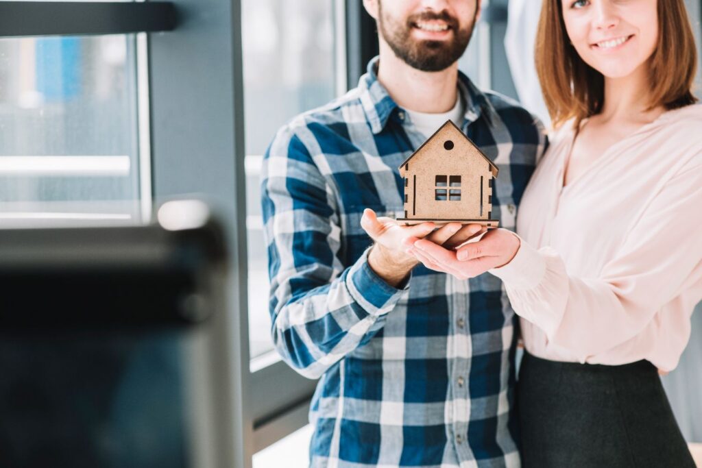 Smiling couple holding a small wooden house model, symbolising buying their first home.