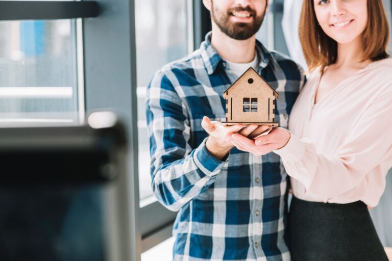 Smiling couple holding a small wooden house model, symbolising buying their first home.